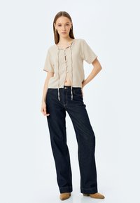 Camisa de botón recortada beige con lazos en el frente, mangas cortas y un ajuste relajado, combinada con jeans de pierna ancha azul oscuro y zapatos beige.