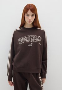 Sudadera marrón con el gráfico "BROOKLYN" en blanco, que presenta acentos a rayas en las mangas y un corte relajado. Material suave de mezcla de algodón.