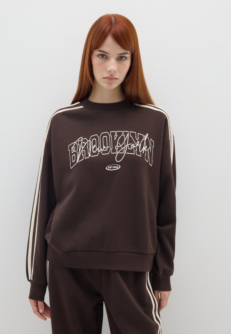 Sudadera marrón con el gráfico "BROOKLYN" en blanco, que presenta acentos a rayas en las mangas y un corte relajado. Material suave de mezcla de algodón.