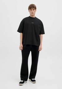 Schwarzes over-sized T-Shirt mit kurzen Ärmeln, rundem Hals und subtiler Logo. Kombiniert mit lockeren schwarzen Hosen und schwarzen Sneakern mit weißen Akzenten.