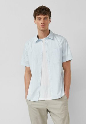 Junger Mann mit kurzen braunen Haaren, der ein hellblaues kurzärmliges Hemd über einem weißen T-Shirt und beigefarbenen Hosen trägt, die Hände in den Taschen.