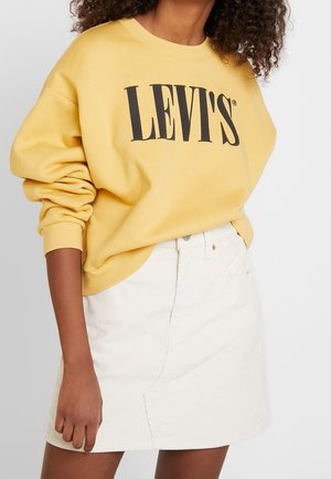Femme portant un sweat jaune Levi's avec un logo noir et une jupe blanche en velours côtelé sur un fond uni.
