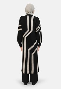 Long black cardigan with vertical cream stripes, made of textured knit material. Features loose sleeves and a straight cut, worn over black pants.