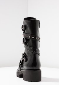 Czarny skórzany botek za kostkę z wieloma paskami, srebrnymi ćwiekami i dodatkami. Gruba podeszwa i klamry dodają teksturowane detale. Gładkie wykończenie.