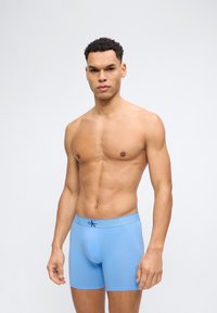 Muskuløs mann iført lyseblå Calvin Klein boxer briefs som står mot en enkel lys bakgrunn, vendt fremover med et nøytralt uttrykk.