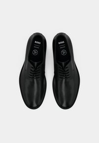 Un paio di scarpe formali in pelle nera con lacci, marchiate "BOSS", esposte affiancate su uno sfondo bianco.