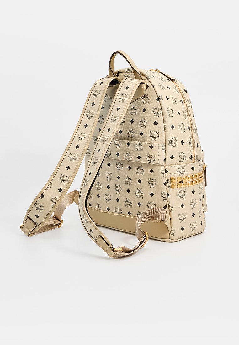 Beige Rucksack mit Logo-Muster, strukturiertem Material, goldenen Hardware-Details, verstellbaren Trägern und einer seitlichen Reißverschlusstasche.