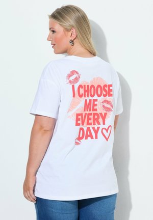 Femme aux cheveux blonds portant un t-shirt blanc avec des empreintes de lèvres rouges et le texte "I choose me every day", debout contre un mur blanc.
