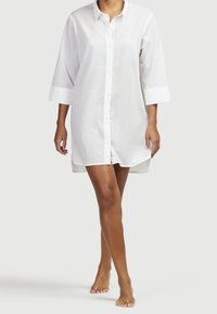 Weißes Hemdblusenkleid mit Dreiviertelärmeln, leicht geschwungenem Saum und leichtem Stoff, getragen von einer stehenden Person.