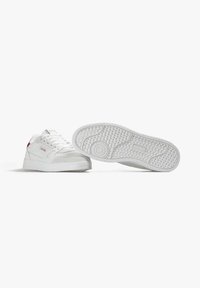 White sneakers with a textured rubber sole, padded tongue, and subtle red detailing. Made of leather and suede with a low-top design.