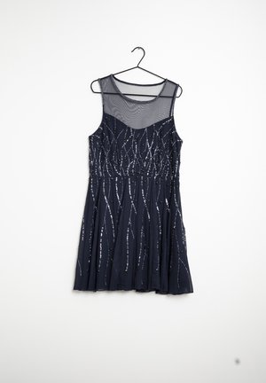 Vestido de cóctel negro sin mangas con escote de malla transparente y patrones lineales de lentejuelas plateadas, colgado en una percha negra sobre un fondo blanco.