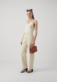 Ljus krämfärgad linne med spetskant, högmidjade beige byxor, svarta remsandaler med klack och en brun läderclutch med texturerad yta.