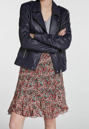Person wearing a black leather jacket over a gray top and a knee-length floral skirt with red and green flowers, arms crossed.