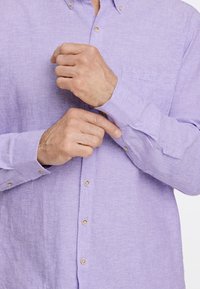 Camisa de lino de color lila claro con un pequeño bolsillo, cuello abotonado y puños abotonados, que presenta una textura suave y un sutil patrón de tejido.
