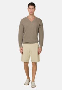 Homme portant un pull col en V taupe, un short beige au genou et des chaussures blanches sans lacets, debout devant un fond blanc.