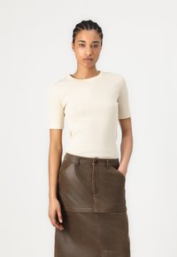 Béžový žebrovaný top s krátkým rukávem spojený s hnědou koženou sukní s knoflíkovým zapínáním a bočními kapsami. Hladká textura po celé ploše.