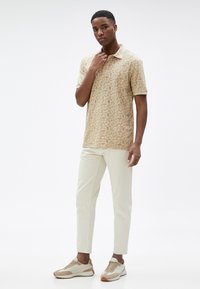 Camisa de polo crema con patrón floral, pantalones beige y zapatillas ligeras. La camisa tiene un cuello y mangas cortas.