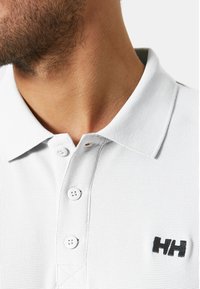 Polo shirt bianco, con texture, dotato di colletto con bottoni, che presenta due bottoni bianchi e un logo nero sul petto. Vista ravvicinata.