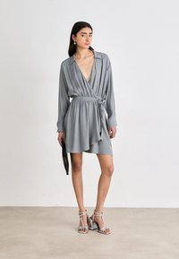 Robe portefeuille grise avec des manches longues, décolleté en V profond, taille cintrée avec lien et ourlet superposé. Associée à des sandales à talons argentées et tenant une pochette noire.