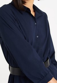 Camisa azul marino de botones con cuello y mangas largas, que presenta un cinturón negro en la cintura y un tejido liso y ligero.
