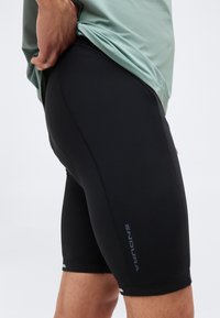 Άτομο που φοράει μαύρο ποδηλατικό σορτς Endura και πράσινο πουκάμισο, κρατώντας το πουκάμισο ψηλά για να δείξει τη γραμμή και το σχεδιασμό του σορτς.