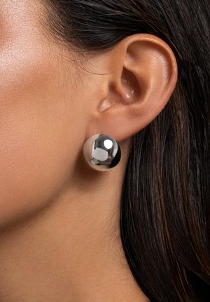 Metallisch silberne Ballohrringe, glatte Oberfläche, kugelförmige Form, große Größe, vor einem strukturierten Hauthintergrund und dunklem Haar.