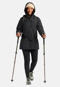 Zwarte waterdichte jas met een capuchon, aangesnoerde taille en zakken. Draag het over zwarte broek en wandelschoenen, geaccessorieerd met wandelstokken.