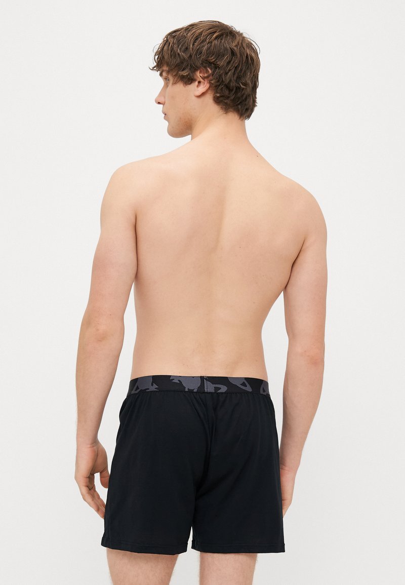 Schwarze Boxershorts mit einem gemusterten grauen Gummibund, aus weichem Stoff. Die Rückansicht zeigt einen lockeren Schnitt und ein knielanges Design.