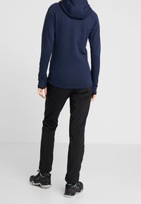 Navyblauer Kapuzenpullover mit langen Ärmeln und Daumenlöchern, kombiniert mit schwarzen Hosen und schwarzen Wanderschuhen. Glatter Stoff, athletischer Schnitt.