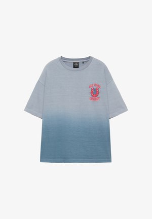 Ein junger Mann steht vor einer beigen Wand, trägt ein blau verlaufendes T-Shirt, beige Shorts und schwarze Flip-Flops mit einer Hand in der Tasche.