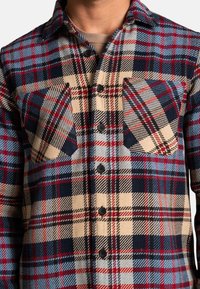 Koszula flanelowa w kratę w niebiesko-czerwono-beżowym schemacie kolorów, z dwiema kieszeniami na piersi, czarnymi guzikami i miękką teksturą.