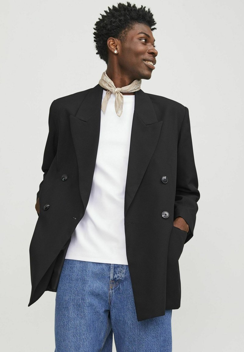 Zwarte dubbele-breasted blazer met grote revers en twee voorzakken, gedragen over een witte T-shirt en gecombineerd met blauwe jeans. Gladde stoftextuur.