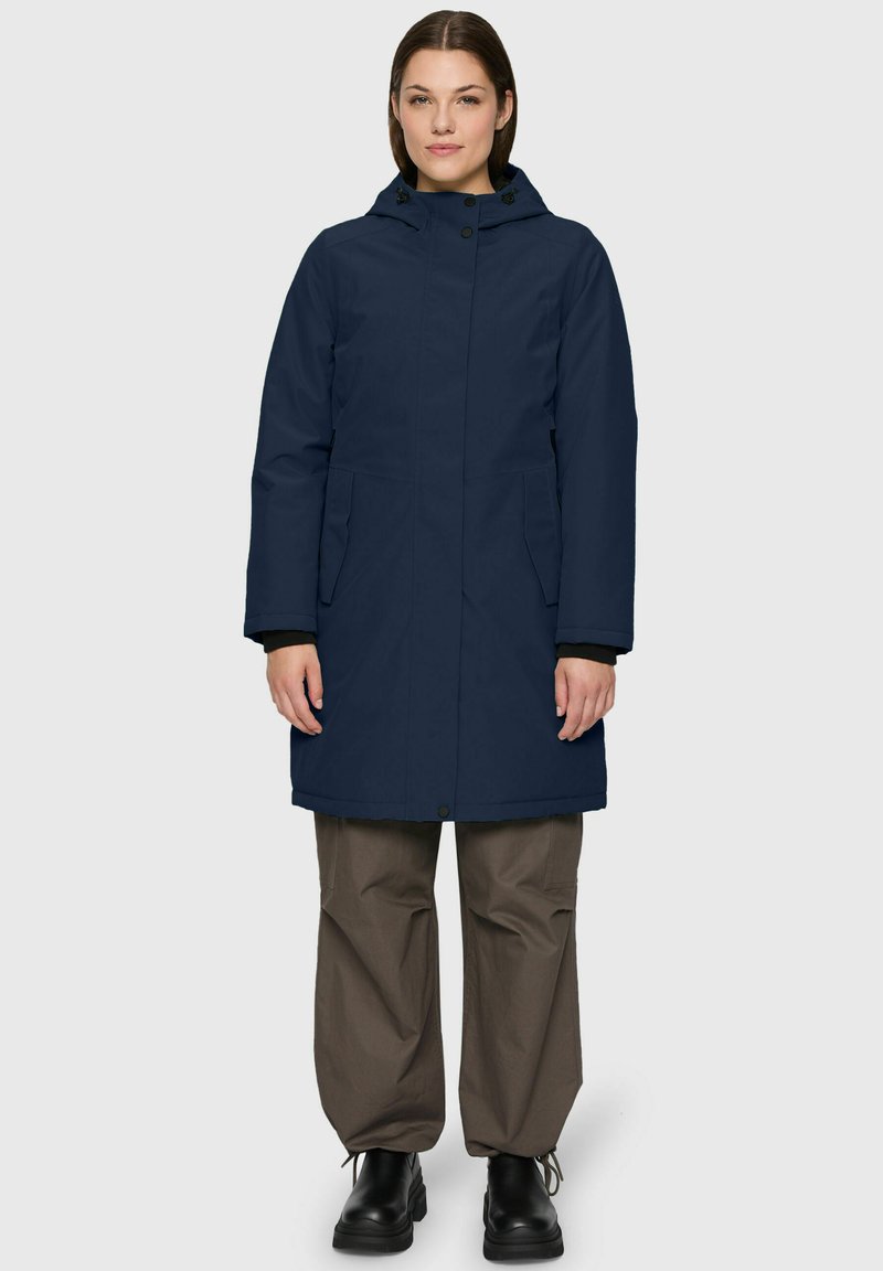 Woman standing facing forward wearing a navy hooded winter coat, brown wide-leg pants, and black lace-up boots.