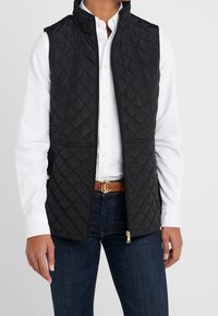 Person som bär en svart vadderad väst över en vit skjorta med knappar, mörkblå jeans och ett brunt bälte med en guldfärgad ankare-spänne.