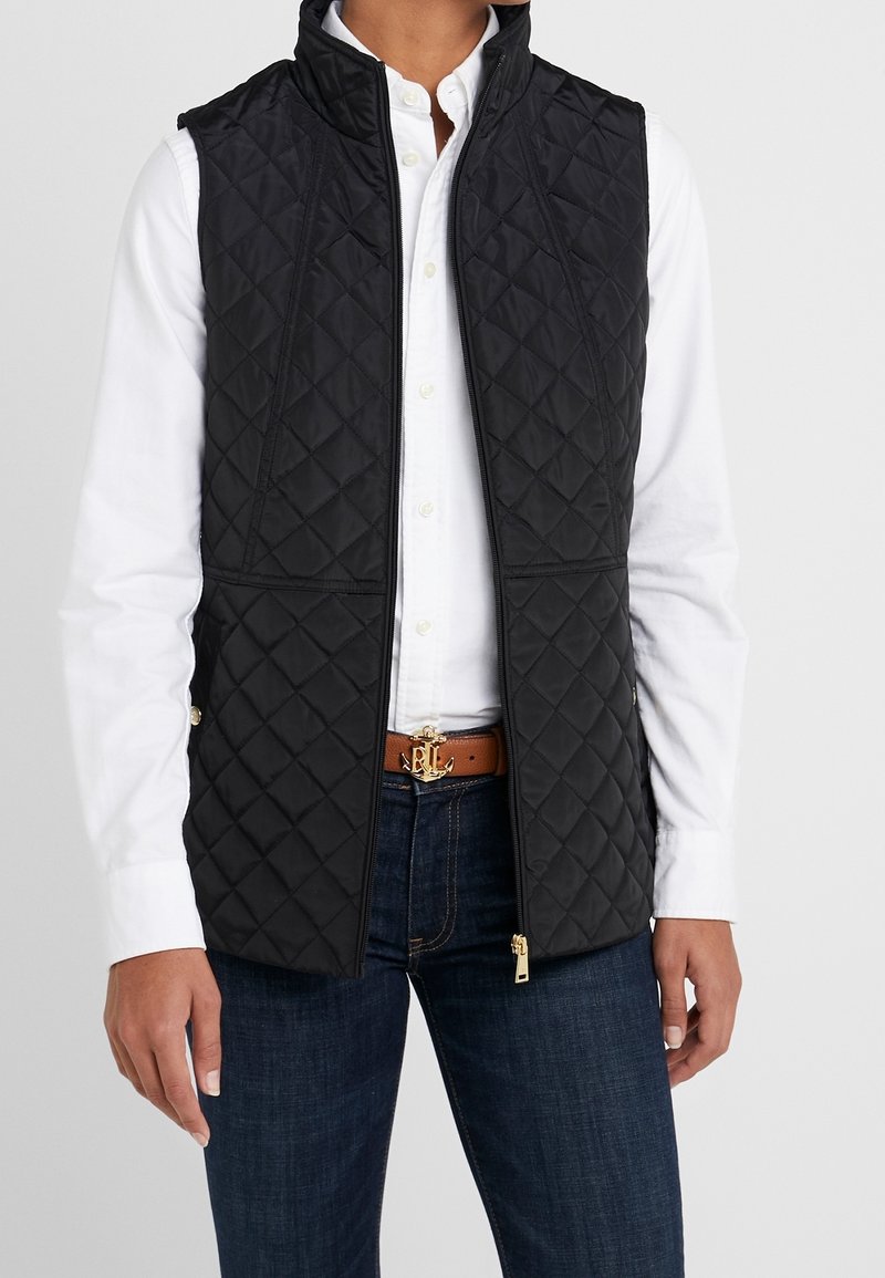 Person som bär en svart vadderad väst över en vit skjorta med knappar, mörkblå jeans och ett brunt bälte med en guldfärgad ankare-spänne.