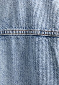 Ljust blå denimtyg med en texturerad väv. Synlig sömnad längs sömmen som visar en standardpassform och denimfinish.