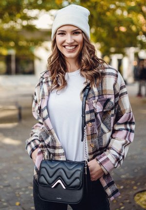 Schwarze gesteppte Handtasche mit einem dekorativen Metallschmuck, kombiniert mit einem mehrfarbigen Karobluse über einem weißen T-Shirt und dunklen Jeans.