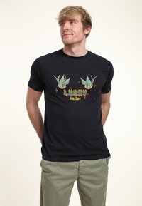 T-shirt noir à manches courtes arborant un graphique coloré « Marin Chanceux » avec deux oiseaux et des étoiles, fabriqué en tissu doux avec une coupe ajustée.
