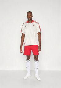 Bílé dresy s tenkými proužky a červenými detaily, červené šortky, bílé ponožky s černými pruhy a bílé kopačky. Sportovní design, atletický střih.