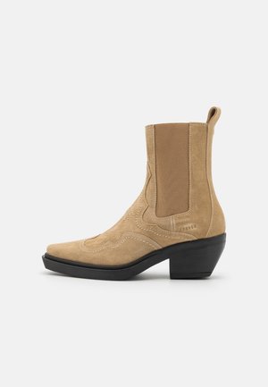 Bottine beige en daim avec talon épais noir, panneau latéral élastique, languette de tirage et coutures décoratives sur la tige.
