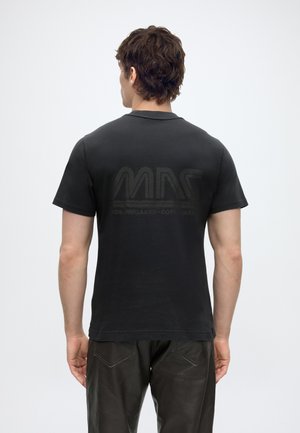 Mies, jolla musta lyhythihainen t-paita, jossa selässä hillitty tumma logo, ja tummat housut, seisoo selin yksivärisen taustan edessä.