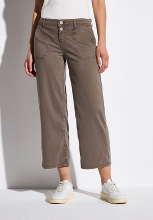 Vrouw draagt cropped wijd uitlopende bruine broek met steekzakken aan de voorkant, dubbele knoopsluiting, witte sneakers en een wit topje tegen een witte achtergrond.