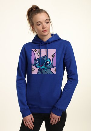 Ung kvinde med blondt hår sat op i en ponyhale iført en blå hættetrøje med en tegneseriefigur med store øjne og stjerner på en lilla baggrund.