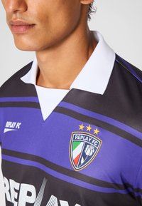 Sort og lilla sportstrøje med en hvid krave, der har et stort "REPLAY FC" logo og tre guldstjerner ovenover.