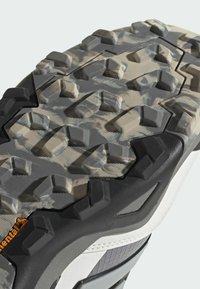 Trail shoe outsole featuring a textured, gray rubber tread with varying shapes and sizes for traction, accented by a black and white midsole.