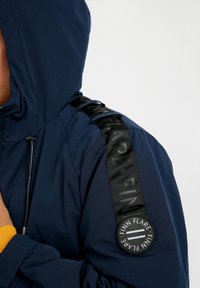 Námořnická kapucová bunda z texturované látky, s černým páskem s logem a kulatou nášivkou na rukávu. Manžety jsou žebrované žluté.