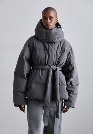 Veste grise duveteuse avec un col montant, une ceinture nouée et un tissu texturé. Le modèle la porte avec un pantalon gris devant un fond neutre.
