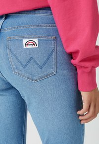 I jeans di denim azzurro chiaro presentano una tasca posteriore con un'etichetta bianca che mostra un logo ad arco rosso e nero, con cuciture e texture visibili.