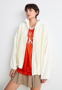 Giacca oversize crema con zip e pieghe verticali, indossata sopra una maglia sportiva rossa. Il modello la abbina a una gonna plissettata beige.