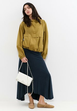 Frau in senfgelber Jacke, dunkelblauem asymmetrischem Rock, beigefarbenen Ballerinas, die eine weiße Tasche mit Kettenriemen hält, steht vor einer weißen Wand.